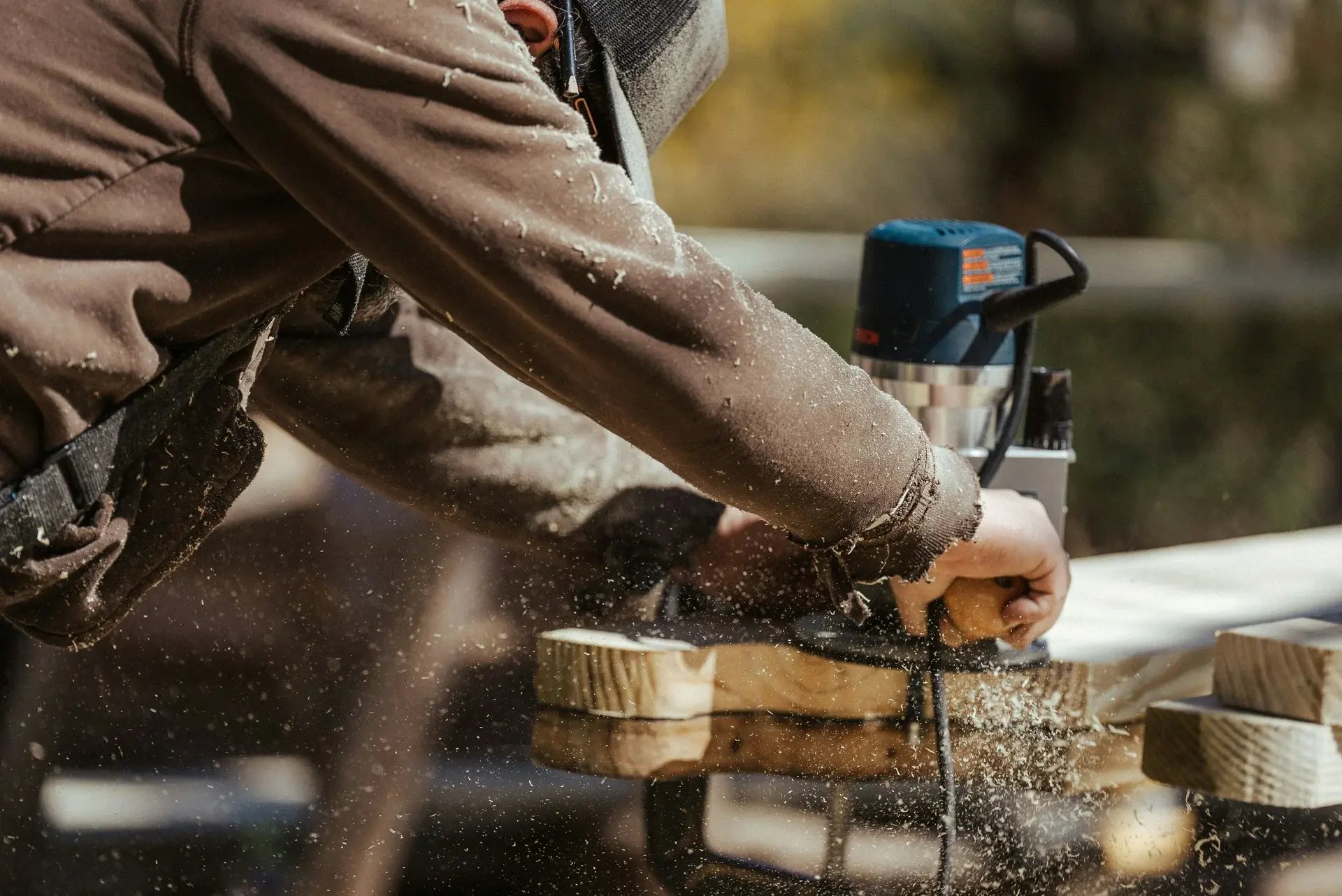 Carpenter using a handheld wood router on lumber, with sawdust and wood shavings flying in sunlight.